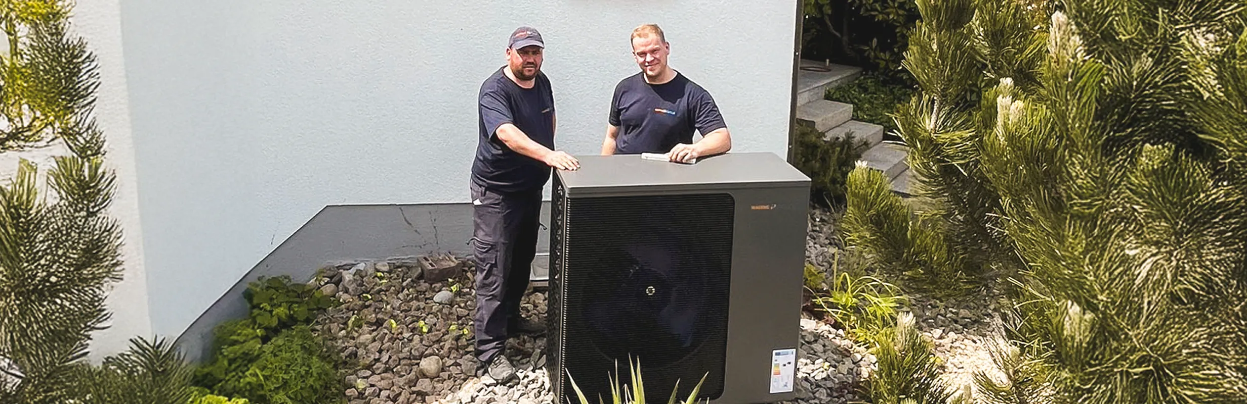 Zwei Teammitglieder des Waermequelle Teams vor einer Waermequelle Wärmepumpe im Garten eines Hauses.