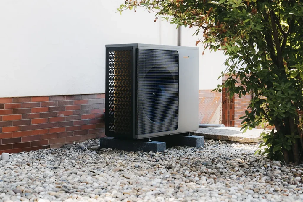 Eine Wärmepumpe der Waermequelle GmbH auf Kieselsteinen vor einer weißen Hauswand.